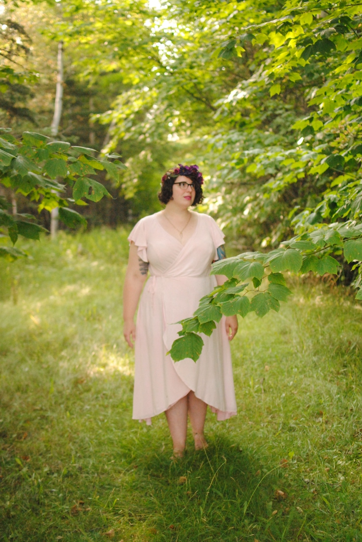 An image of Shannon standing in the woods, surrounded by greenery, wearing a pink wrap dress and a purple floral crown. They look off to the right with a soft expression.