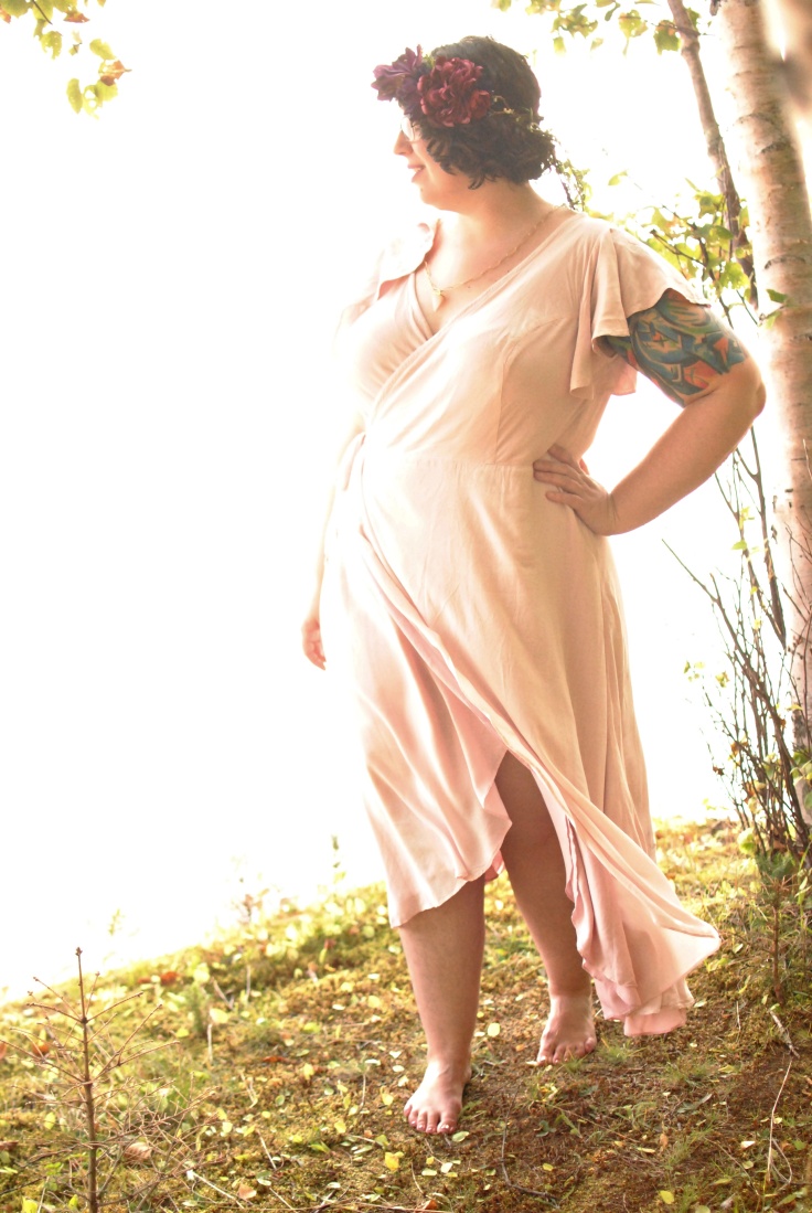 An image of Shannon standing near the edge of a cliff, wearing a pale pink wrap dress that flutters around their legs. The ground is green and mossy and the background is washed out and bright.