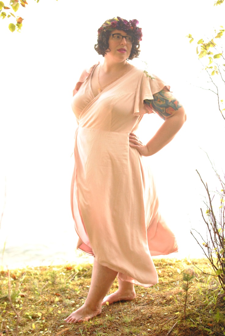 Shannon at the cliff's edge, wearing a pink wrap dress that flutters around her arms and legs in the wind. She twists to look over one shoulder with a hand on her hip. The light is bright and back-light, with a golden cast.