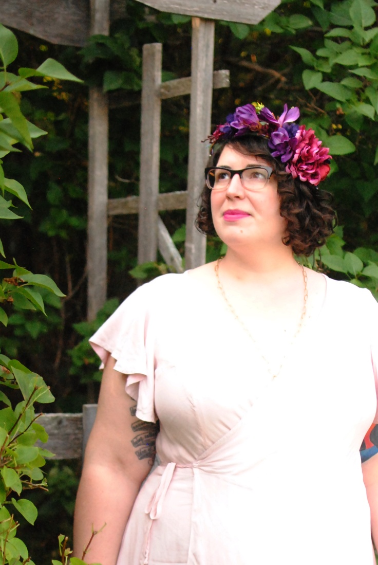 A half-length portrait of Shannon looking up and to one side, with a purple flower crown on top of their curly, dark hair. They have a soft, wistful smile on their face.