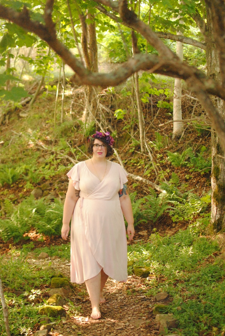 Shannon walking through a wooded area, with dappled golden light falling around them and on the mossy ground. Shannon wears a pink wrap dress and a purple flower crown, and looks at the camera as they walk forward.