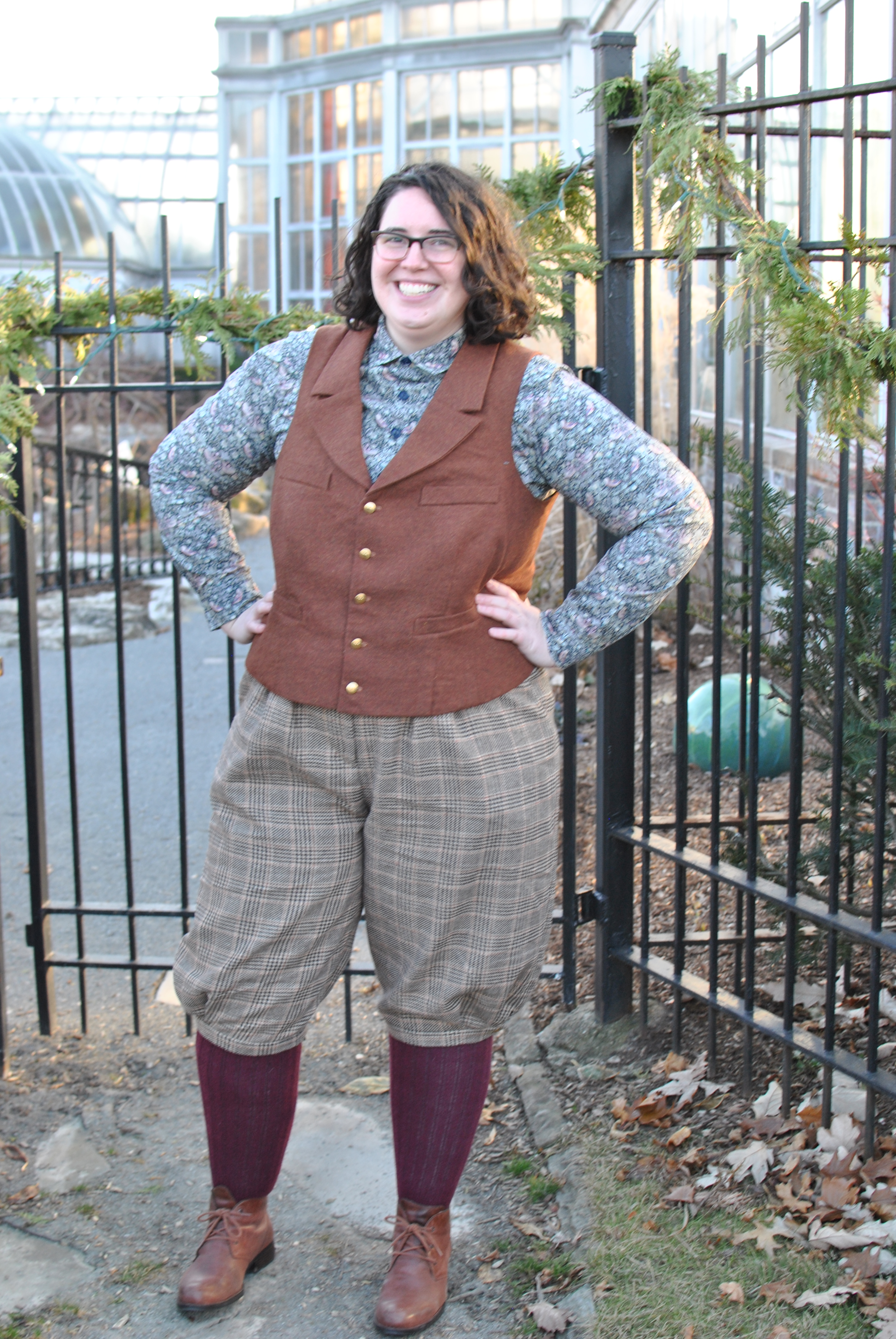 Shannon standing outdoors in front of an iron gate, wearing plaid knickerbockers, a rust wool vest, and a patterned shirt.