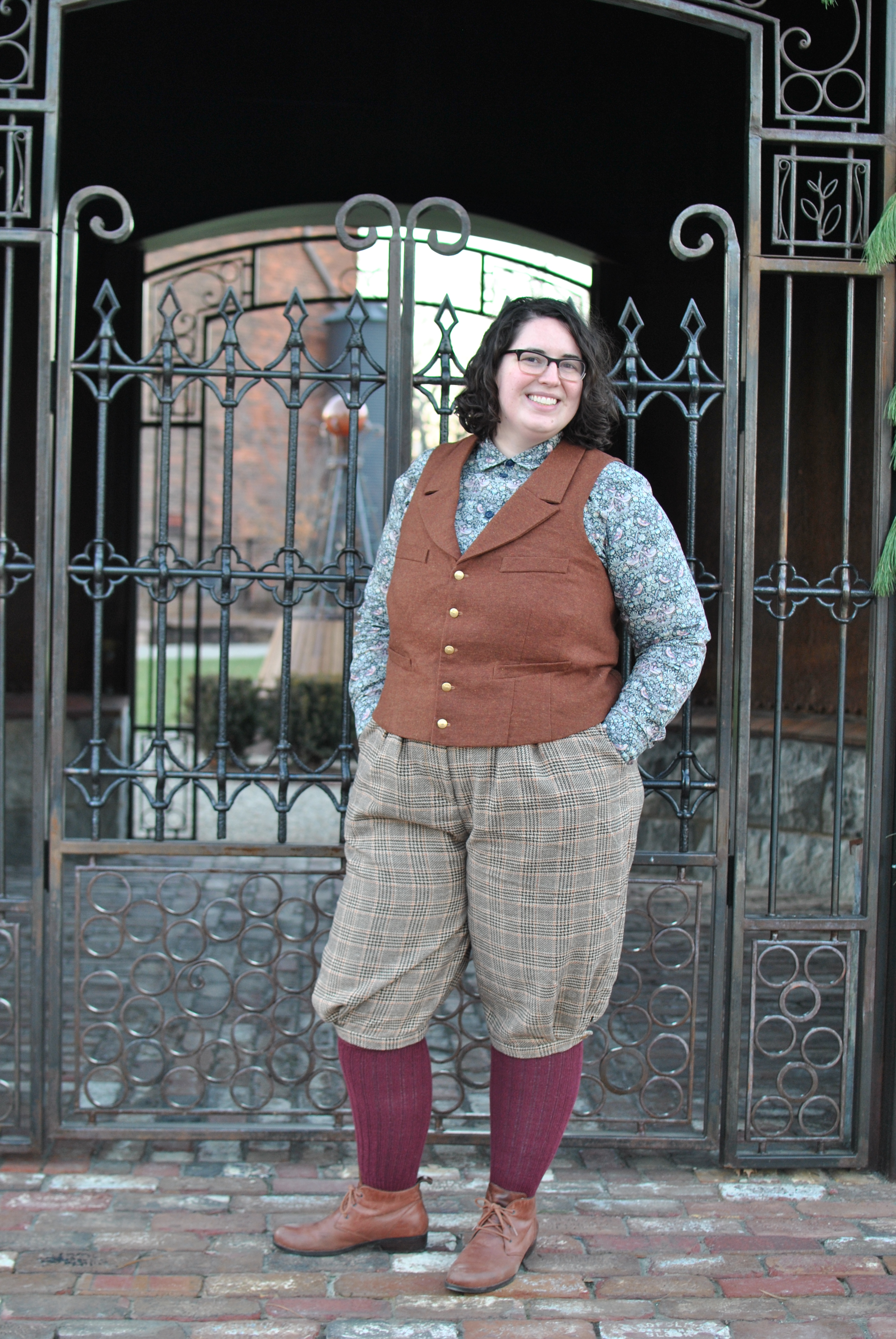 Shannon standing outdoors in front of an iron gate, wearing plaid knickerbockers, a rust wool vest, and a patterned shirt.