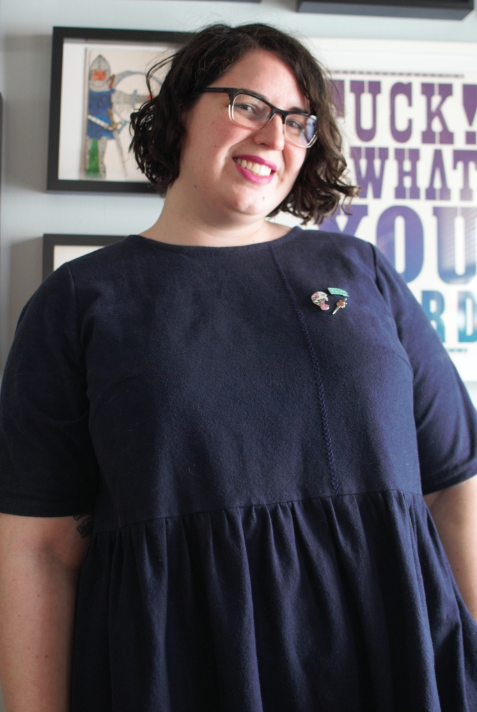 Shannon in a half length portrait, smiling at the camera. More visible here is the line of embroidered stitching down the front of the dress bodice and a little cluster of pins.